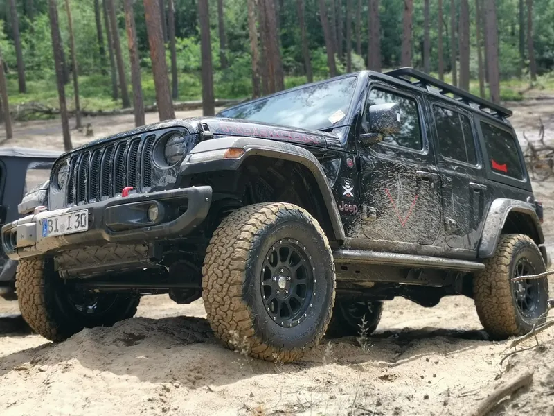Schwarzer Jeep Wrangler mit Schlammspuren auf sandigem Waldboden