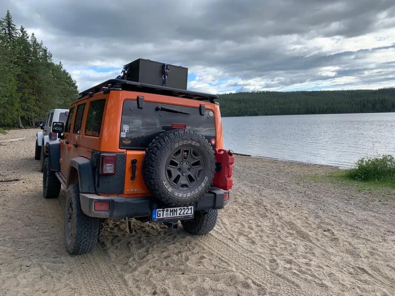 Oranger Jeep Wrangler mit Dachbox am Sandstrand eines Sees