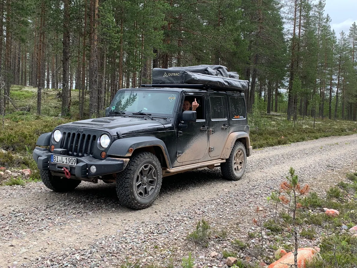 Grauer Jeep Wrangler mit Dachzelt auf einer Schotterstraße im Kiefernwald